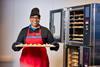 A smiling woman in a Greggs uniform holding a baking tray of sausage rolls