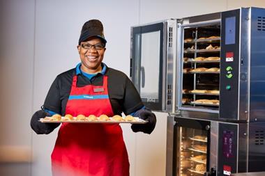 A smiling woman in a Greggs uniform holding a baking tray of sausage rolls