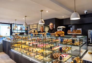 Two smiling people behind a beautifully stocked bakery counter