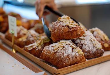 A pair of tongs picking up an almond croissant from a pile