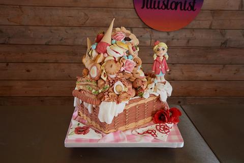 A picnic themed cake with food, including sandwiches, cakes, and meat, piled high in a woven basket