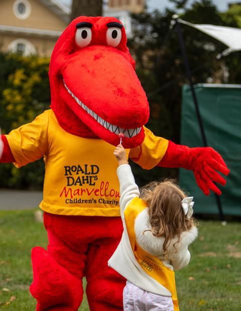A young child offers a lollipop to a red dinosaur character wearing a Roald Dahl’s Marvellous Children’s Charity t-shirt