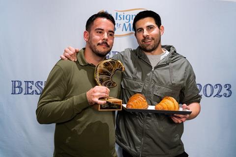 Two dark haired men holding a croissant-shaped trophy and a tray of freshly baked croissants