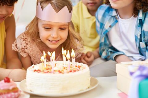A girl wearing a paper crown surrounded by other children prepares to blow out candles on a birthday cake.