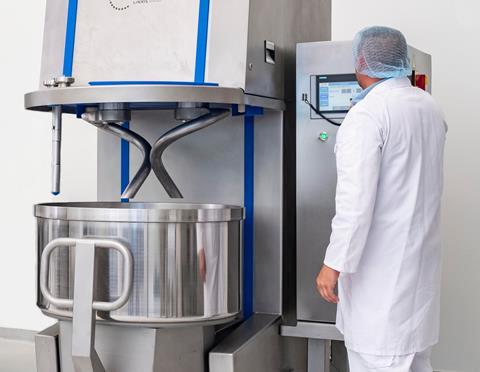 A man in a white coat and hairnet with a vertical mixer and kneader