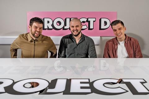 Three men sat at a table with a pink 'Project D' sign behind them