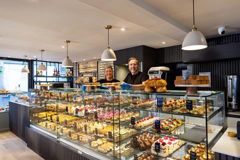 Two smiling people behind a beautifully stocked bakery counter