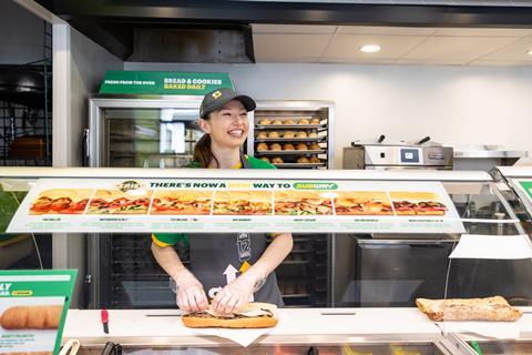 A female staff member smiles while making a Subway sandwich at a counter.