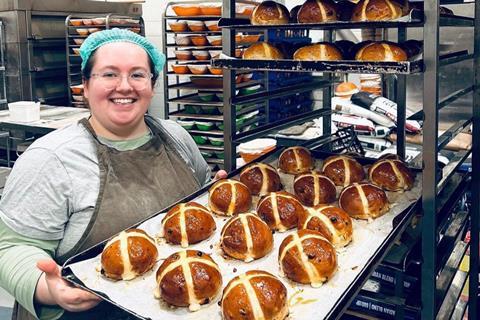 A smiling baker with a hairnet on holding a tray of hot cross buns