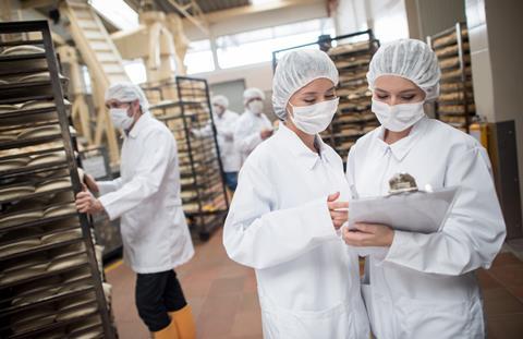 Two women in white outfits, hairnets, and face masks checking for quality at an industrial bakery