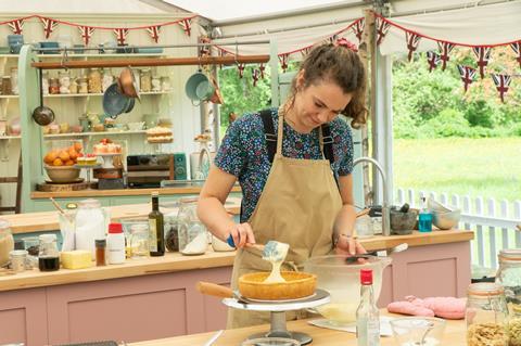 A woman with curly hair and a beige apron in the Bake Off tent putting icing on a biscuit