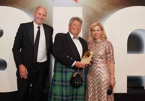 A man in a tuxedo, a man in a kilt and a blonde woman in a sparkly dress at an award ceremony
