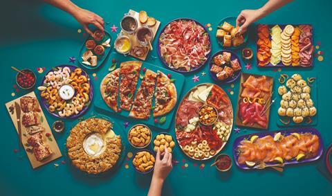 A selection of party food on a blue table including sausage rolls, cheese and crackers, pizza, smoked salmon and more