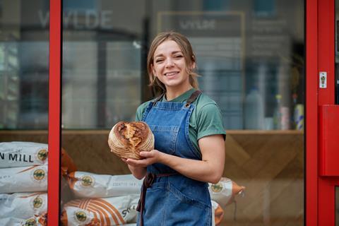 Naomi Spaven in a denim apron proudly holding a loaf of sourdough