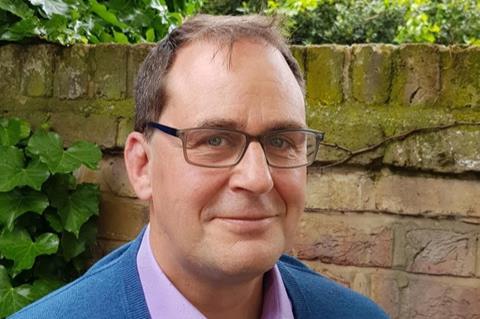 A headshot of Ed Heaver, a man with brown hair and glasses next to a brick wall