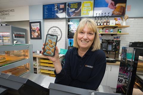 A woman working in a Warrens Bakery store, holding a phone with the Warrens Bakery Rewards app displayed on it