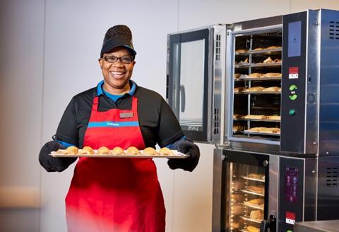 A smiling woman in a Greggs uniform holding a baking tray of sausage rolls