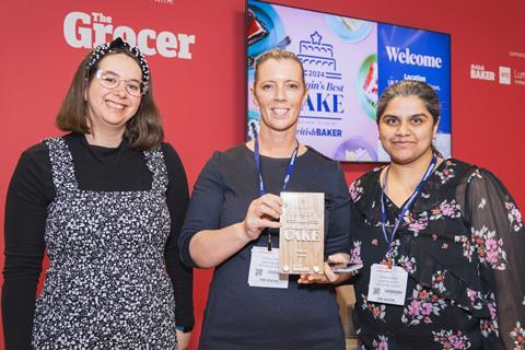 Three women smile with a wooden cake trophy in front of a red background