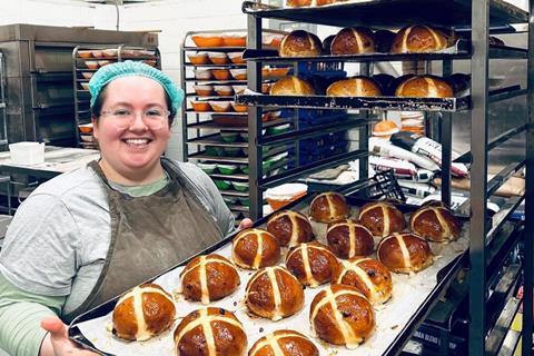 A female baker in a grey apron and hair net holding a tray of hot cross buns