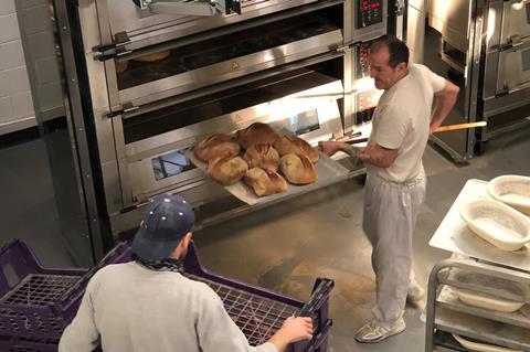 A baker dressed in white clothes taking sourdough loaves out of the oven on a peel