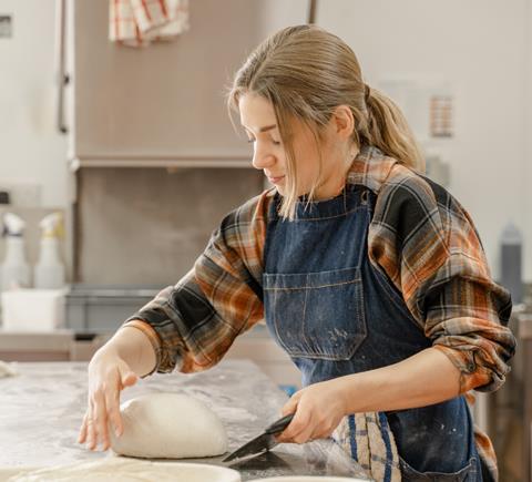 A blonde haired female baker in denim apron shaping bread dough