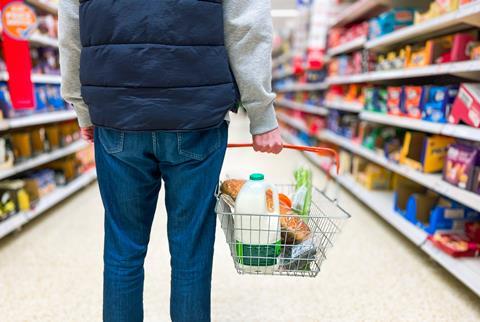 A person in jeans holding a basket with milk, bread and groceries in, standing in the biscuit aisle of a supermarket