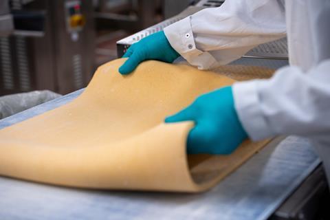 A person in a white jacket and blue gloves handles a sheet of dough