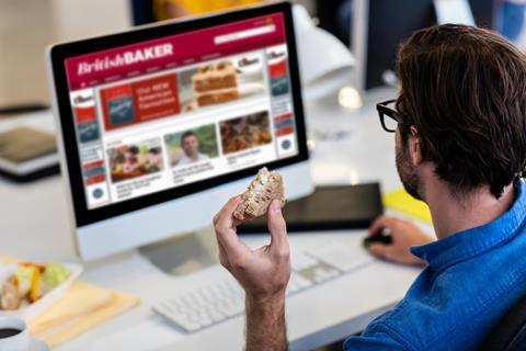 A man eats a sandwich at a work desk while browsing the British Baker website